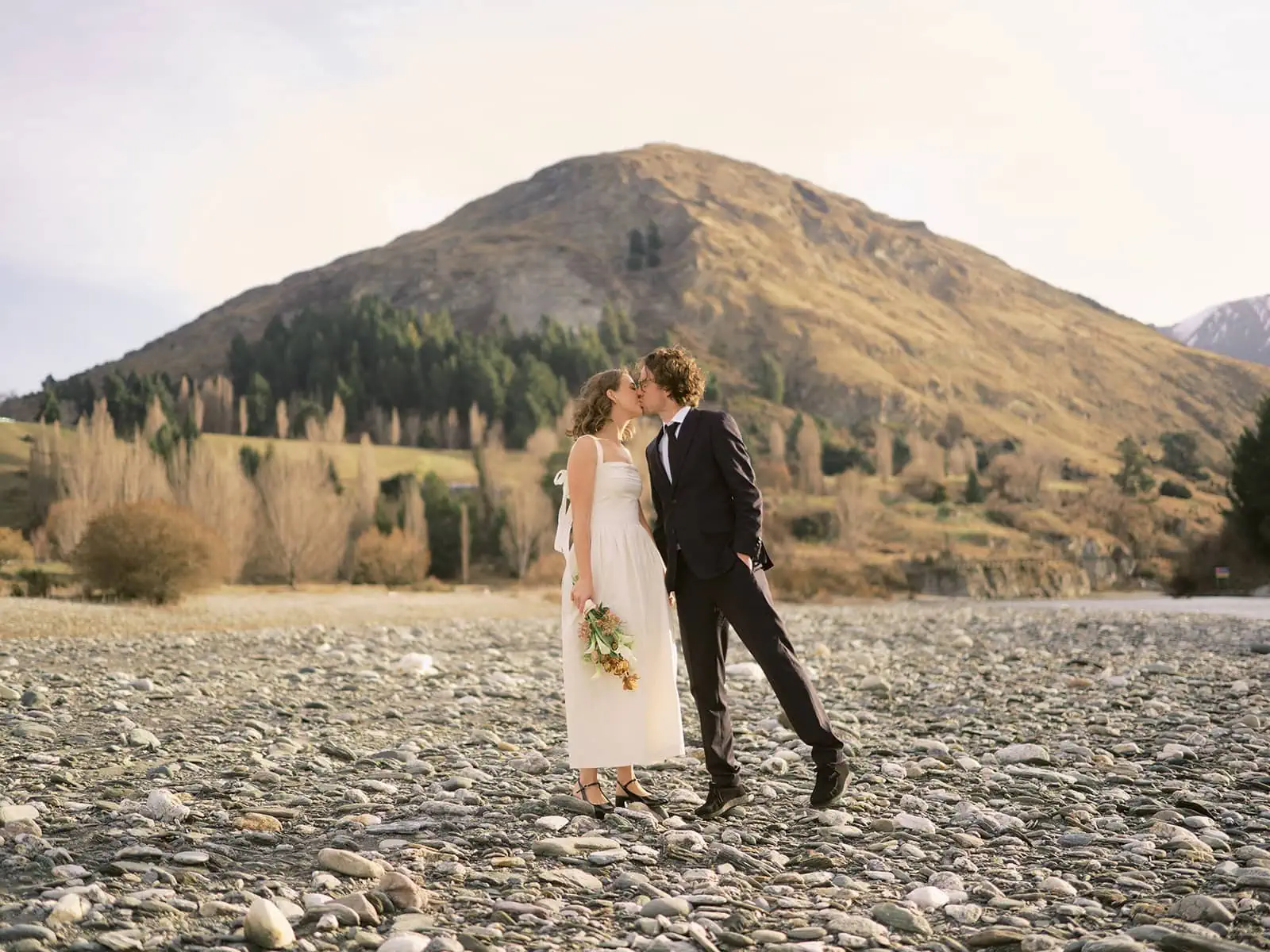 queenstown new zealand heli wedding elopement photographer videographer | A couple dressed in formal attire shares a kiss on a rocky riverbed, capturing a romantic elopement moment against the stunning backdrop of Queenstown's hills and sparse trees, all under a clear sky.