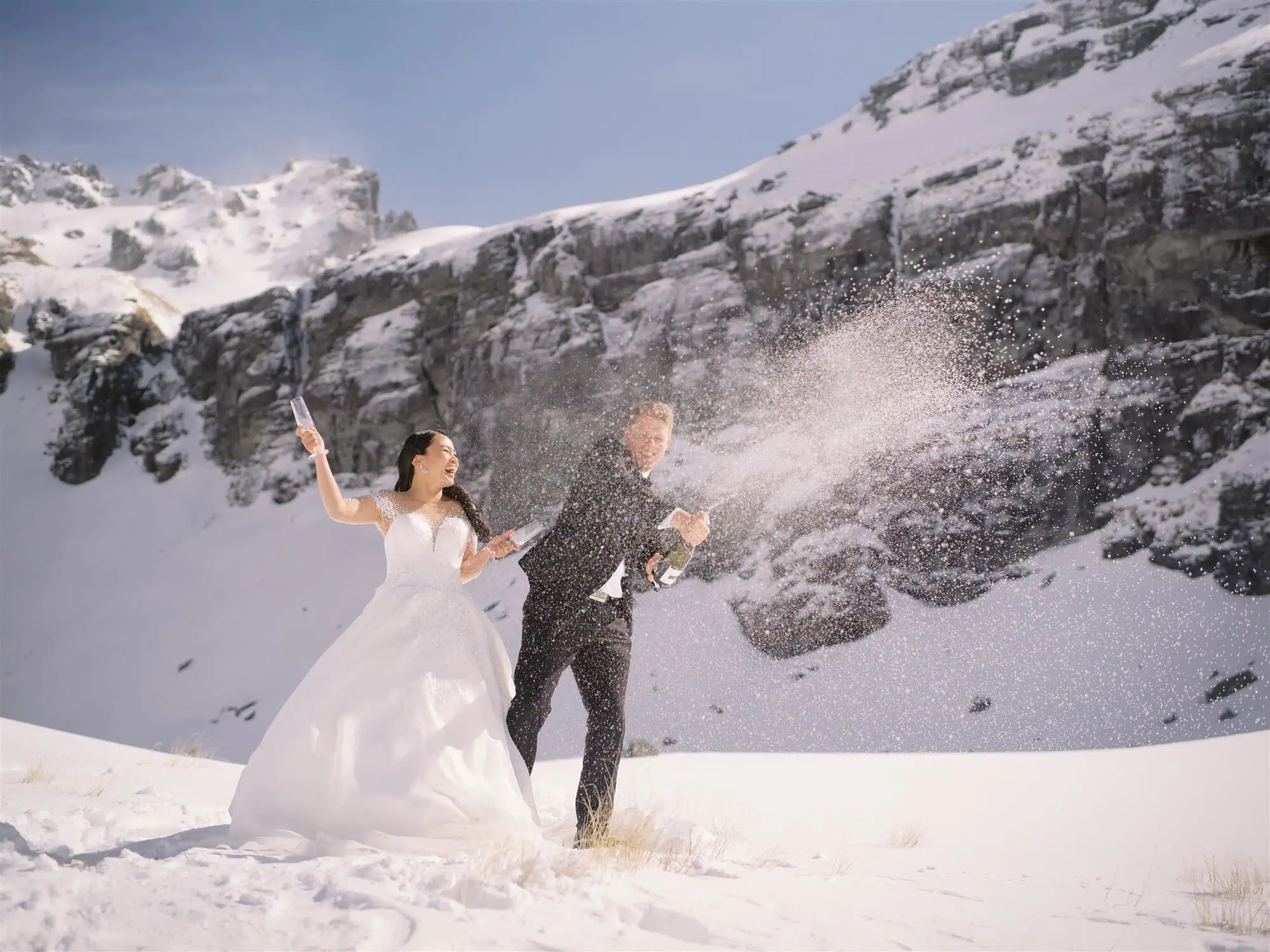 queenstown new zealand heli wedding elopement photographer videographer | In a stunning Queenstown elopement, the bride and groom celebrate outdoors in snowy mountains, spraying champagne with wide smiles.