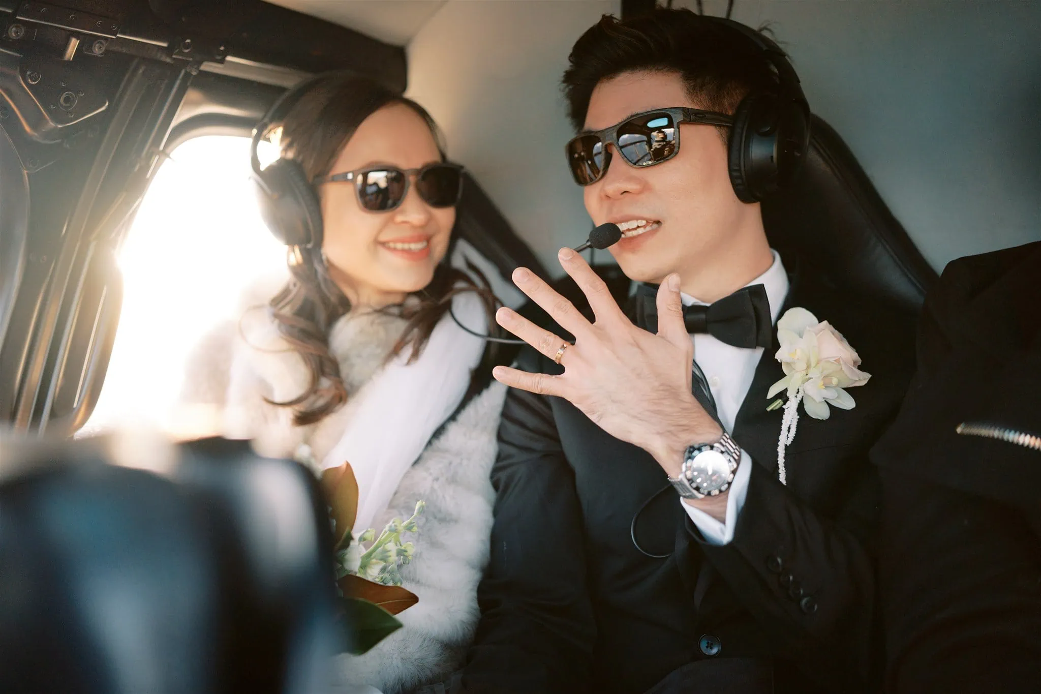 queenstown new zealand heli wedding elopement photographer videographer | Tiffany & Malcolm, dressed in elegant formalwear and headsets, sit inside a helicopter, their smiles capturing the spirit of a Queenstown elopement. The man gestures warmly, adding a touch of candid charm to what feels like an exquisite moment of wedding photography.