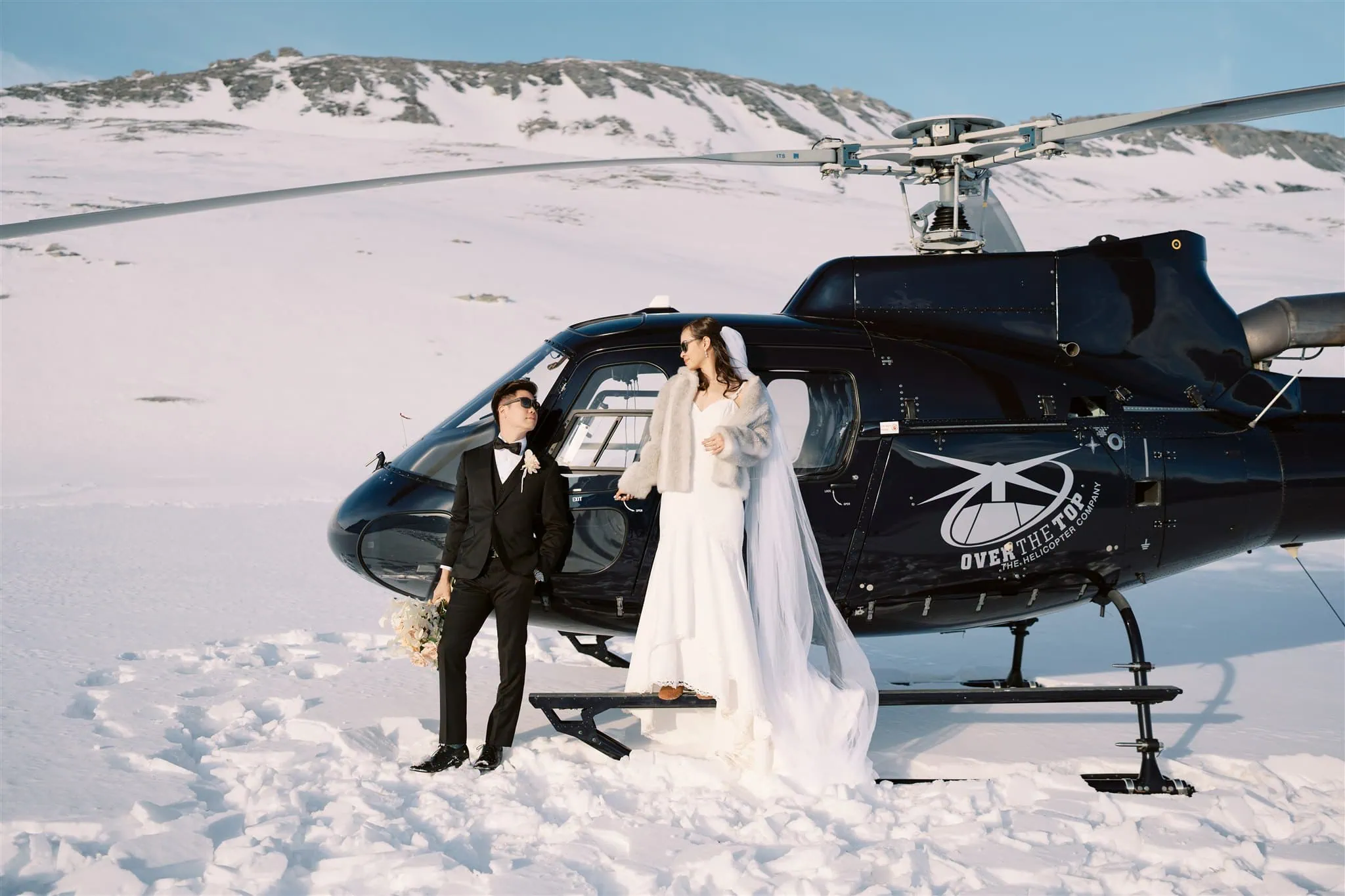 queenstown new zealand heli wedding elopement photographer videographer | A bride and groom in formal attire stand near a black helicopter on a snowy Queenstown landscape. Capturing the essence of elopement, the bride dazzles in her white dress while the groom looks sharp in his black suit—perfect moments for breathtaking wedding photography.