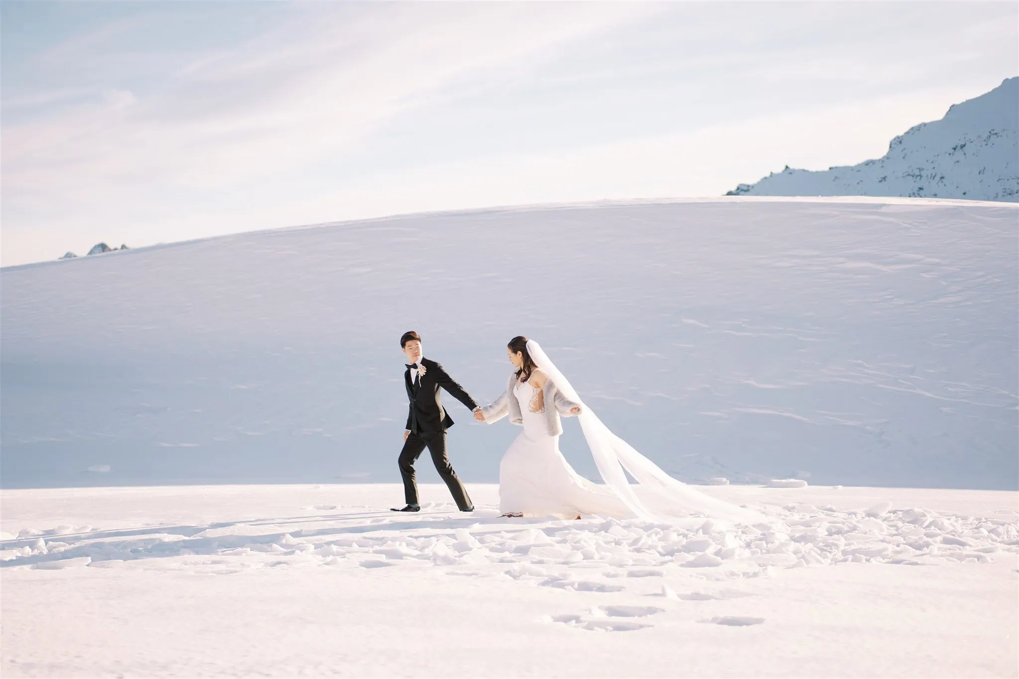 queenstown new zealand heli wedding elopement photographer videographer | Tiffany and Malcolm stroll hand in hand through the snowy landscape of their Queenstown elopement. She dazzles in a white wedding dress with a long veil, while he is elegantly clad in a black tuxedo—captured beautifully for their wedding photography collection.