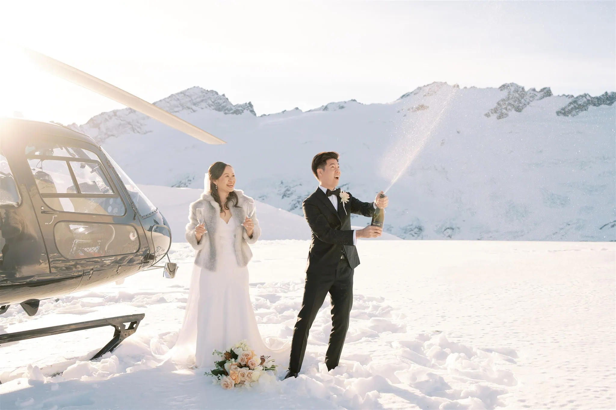 queenstown new zealand heli wedding elopement photographer videographer | Tiffany & Malcolm celebrate their Queenstown elopement with an unforgettable moment. Amidst the snowy mountains, they stand beside a helicopter as the groom joyfully sprays champagne, capturing the essence of wedding photography magic.