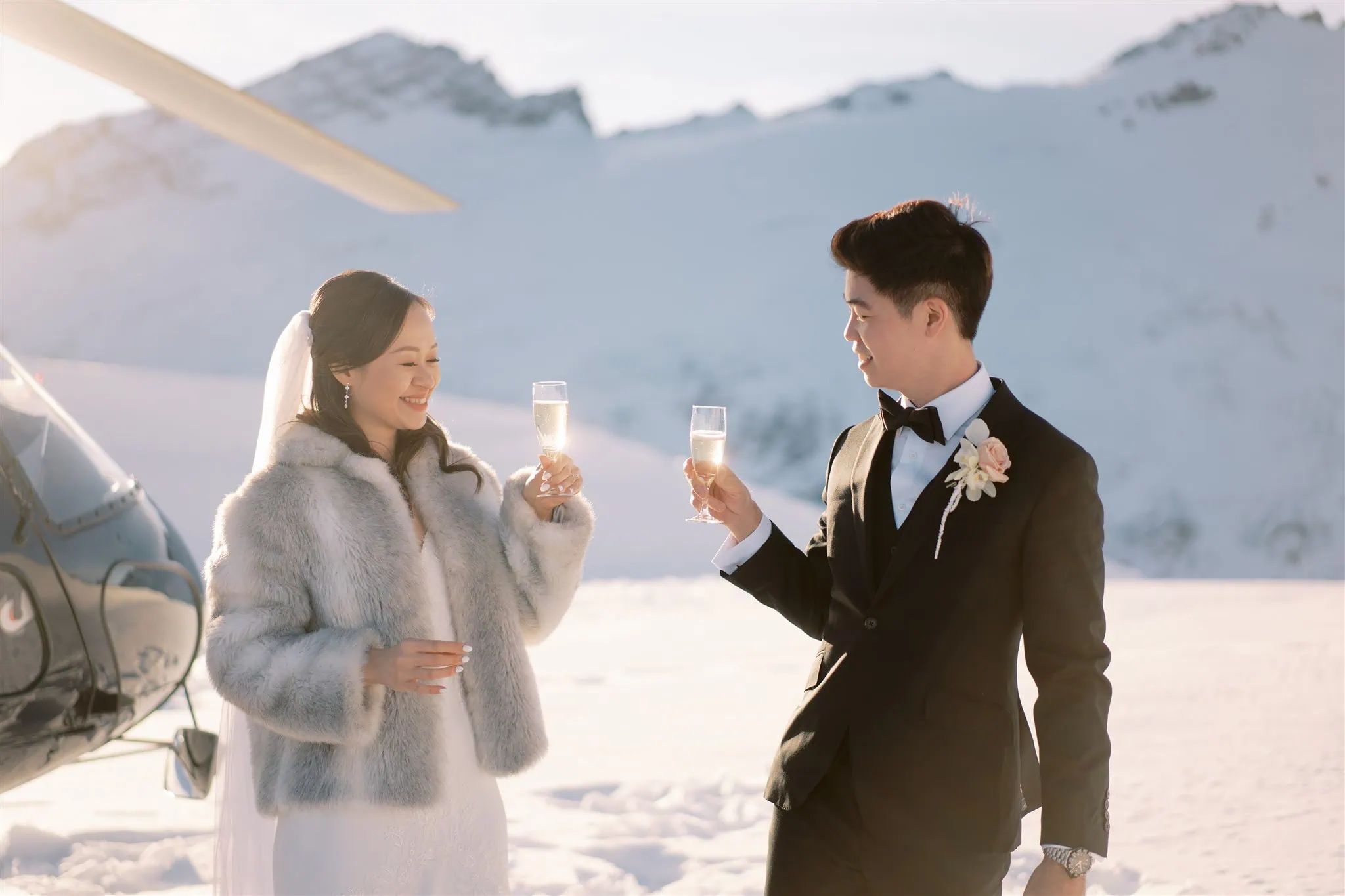 queenstown new zealand heli wedding elopement photographer videographer | A couple in wedding attire clinks glasses of champagne, celebrating their Queenstown elopement in a snowy mountain landscape next to a helicopter—a picture-perfect moment of wedding photography.