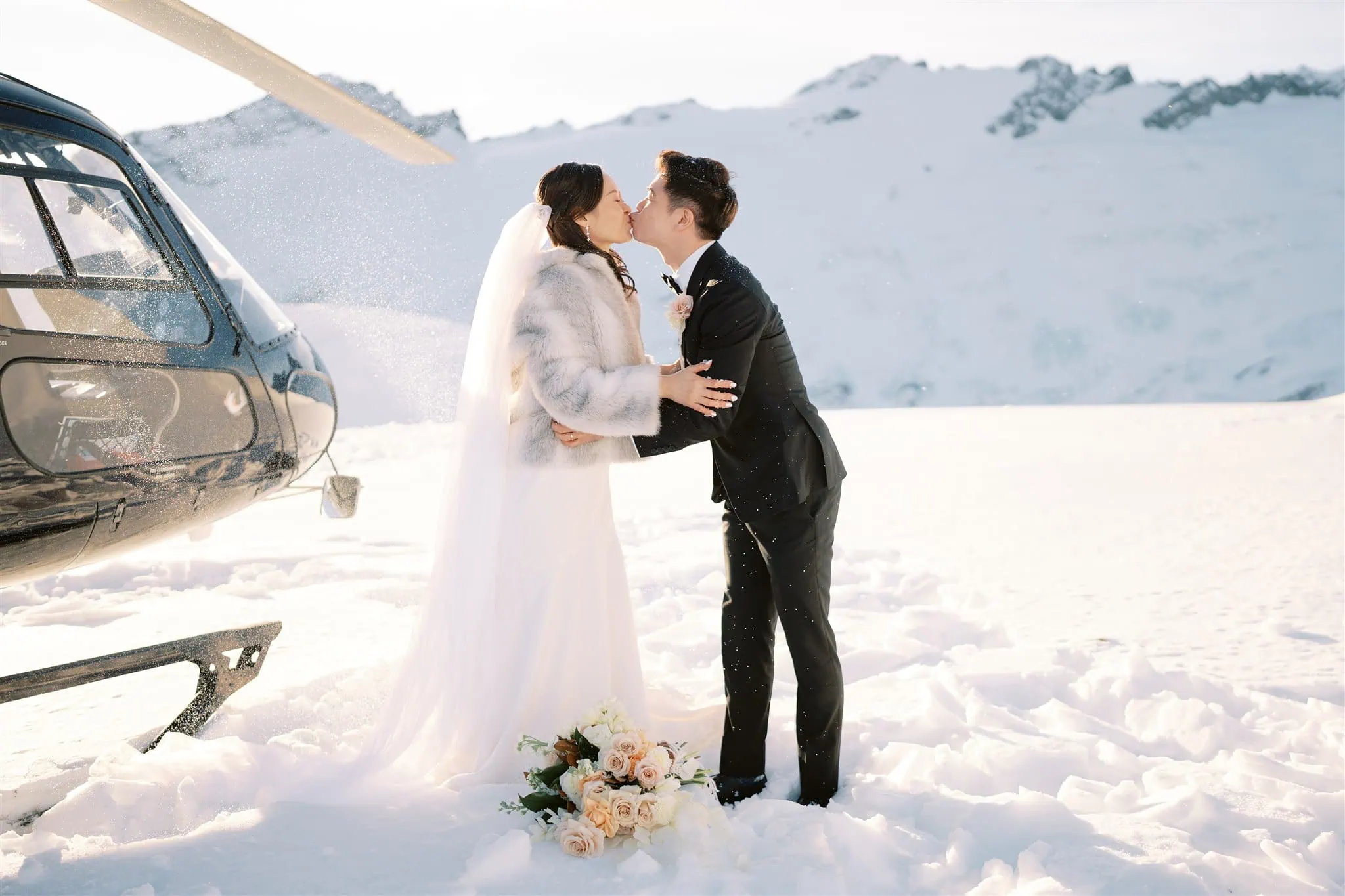 queenstown new zealand heli wedding elopement photographer videographer | Tiffany & Malcolm share a romantic kiss on a snowy Queenstown mountaintop, surrounded by a helicopter and a bouquet of flowers, beautifully captured for their wedding photography.