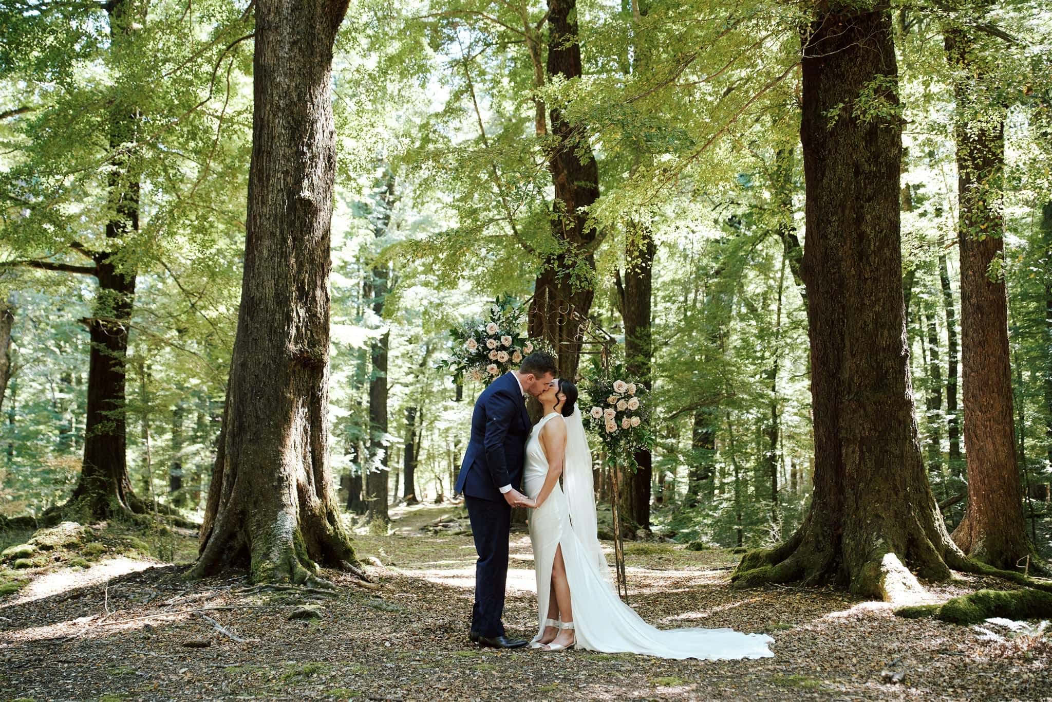 queenstown new zealand heli wedding elopement photographer videographer | Odylia and Ben share a tender kiss amidst the tall trees of a forest, sunlight gently filtering through the leaves during their intimate Glenorchy elopement wedding.
