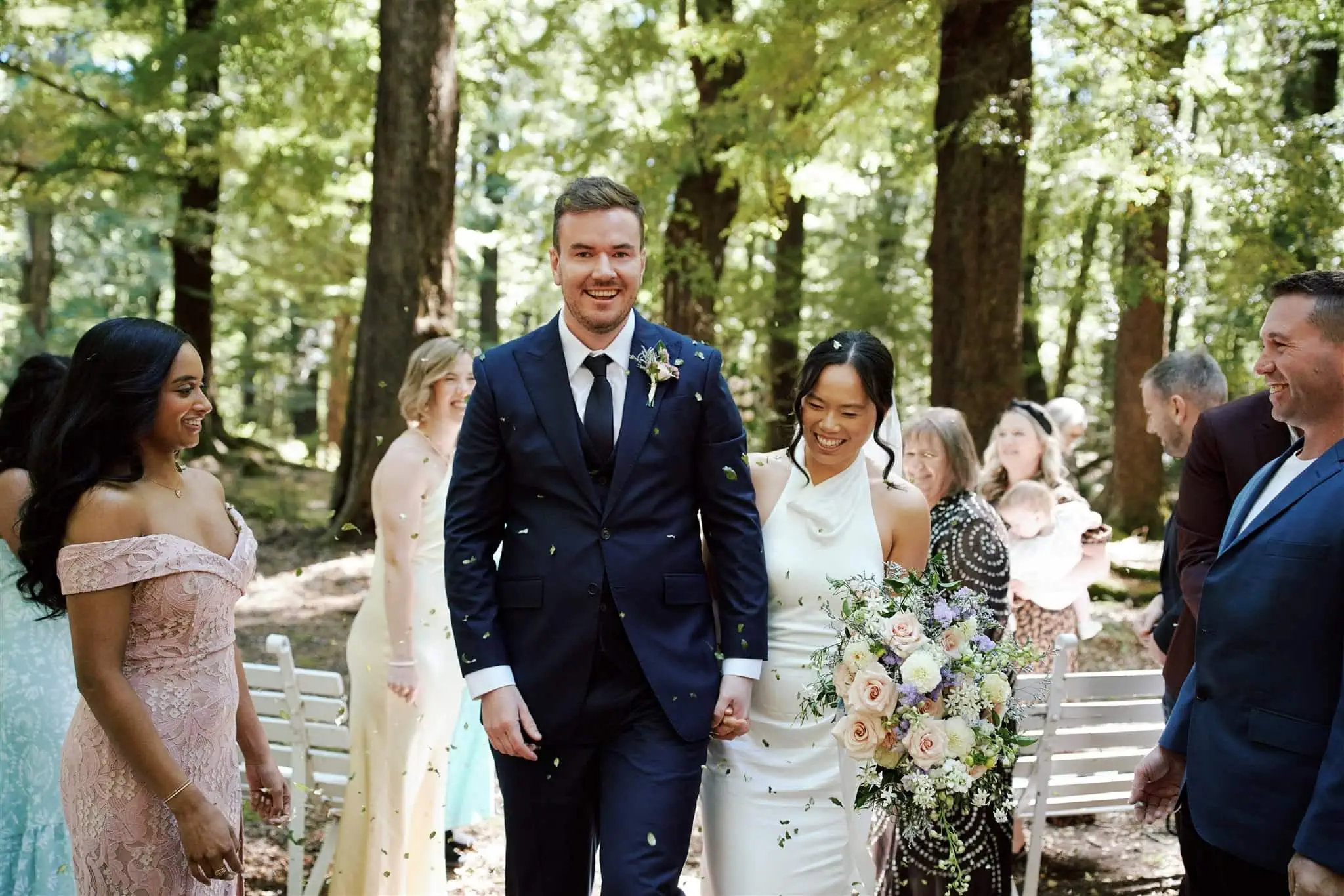 queenstown new zealand heli wedding elopement photographer videographer | Odylia & Ben are all smiles as they walk down the outdoor aisle at their Glenorchy elopement wedding, surrounded by guests in formal attire and framed by a stunning backdrop of trees.