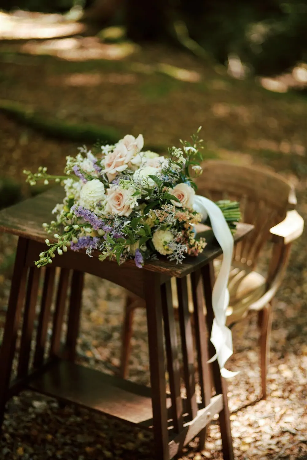 queenstown new zealand heli wedding elopement photographer videographer | A bouquet of white, pink, and purple flowers with a white ribbon rests on a small wooden table in a shaded, wooded area. An empty wooden chair stands nearby, capturing the serene essence of an intimate Glenorchy Elopement Wedding.