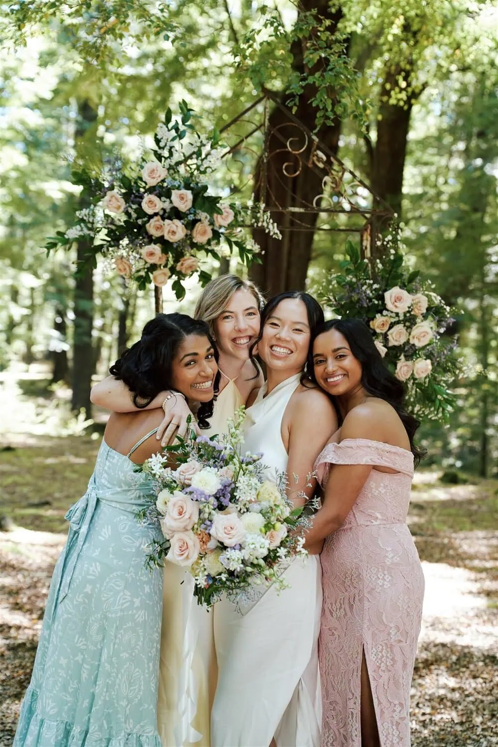 queenstown new zealand heli wedding elopement photographer videographer | Under a decorative flower arch nestled in the woods of Glenorchy, four women in formal attire embrace and smile. Three don pastel dresses while one, likely celebrating an elopement wedding with Odylia & Ben, is clad in a white gown, clutching a vibrant bouquet.