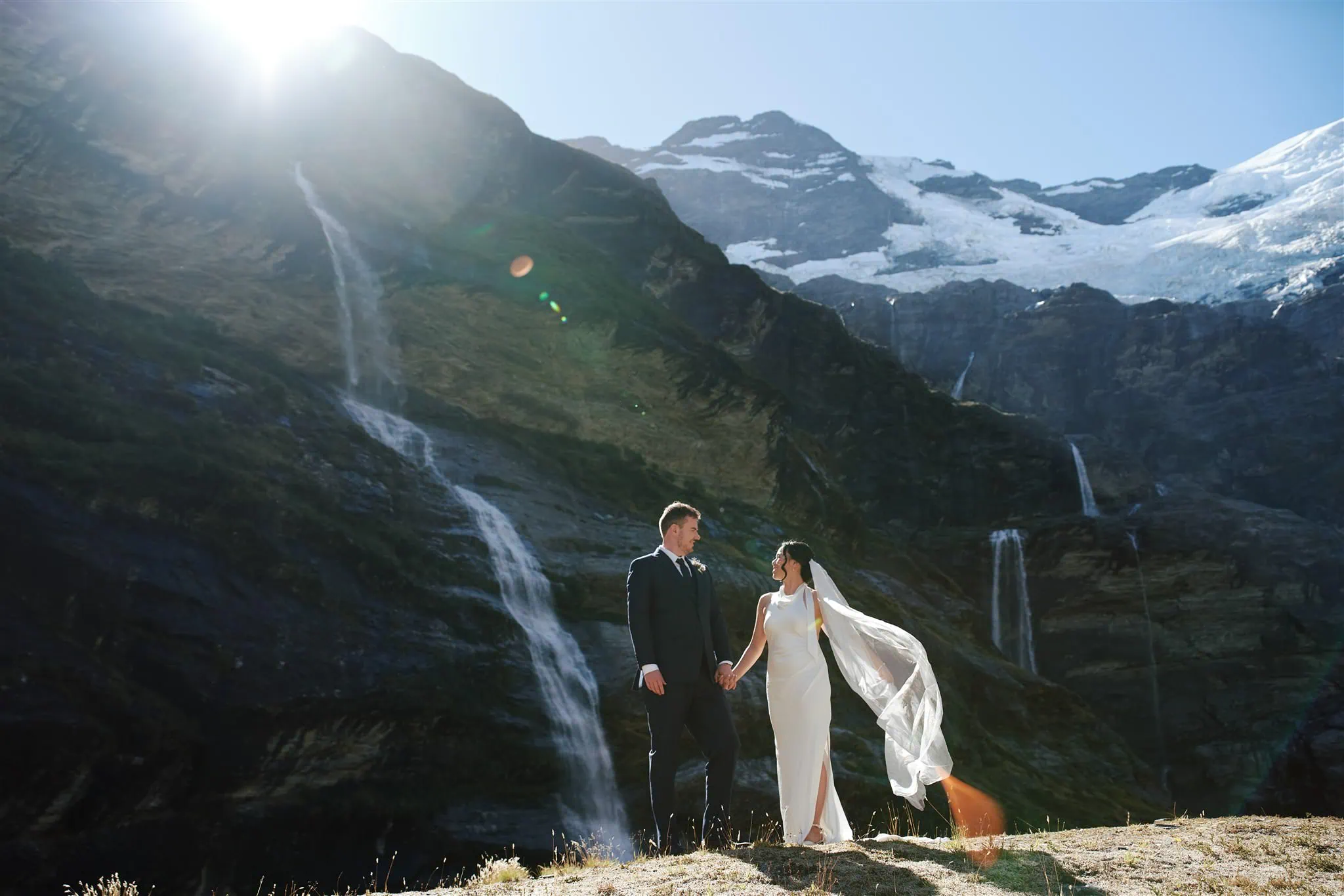 queenstown new zealand heli wedding elopement photographer videographer | Odylia & Ben stand hand in hand on a mountain ledge, celebrating their Glenorchy Elopement Wedding against a breathtaking backdrop of waterfalls and snow-covered peaks under bright sunlight.