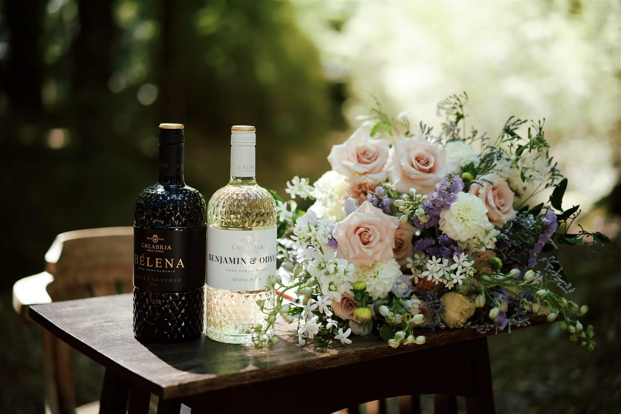 queenstown new zealand heli wedding elopement photographer videographer | Two wine bottles and a floral arrangement grace a wooden table outdoors at an Elopement Wedding in Glenorchy. One bottle is red, the other white, accompanied by pink roses and lush greenery. Odylia & Ben's intimate celebration is perfectly captured in this serene setting.