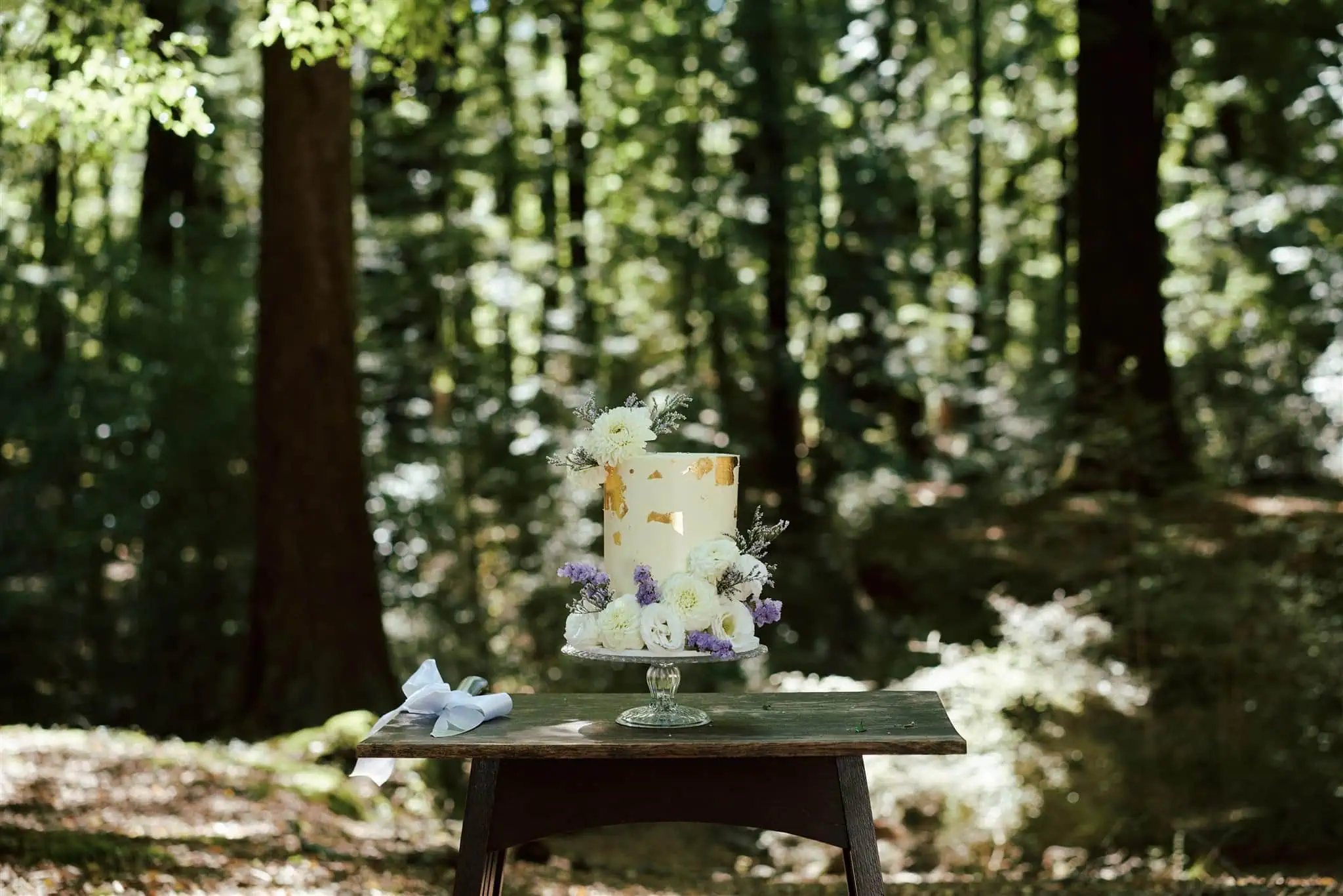 queenstown new zealand heli wedding elopement photographer videographer | A tiered wedding cake adorned with white and purple flowers graces a wooden table in a sunlit forest during Odylia & Ben's Glenorchy Elopement Wedding.