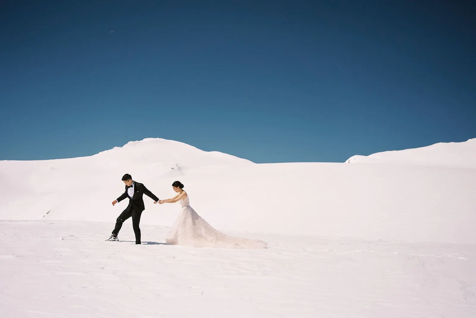 queenstown new zealand heli wedding elopement photographer videographer | A bride and groom walk hand in hand on a snow-covered landscape under a clear blue sky.