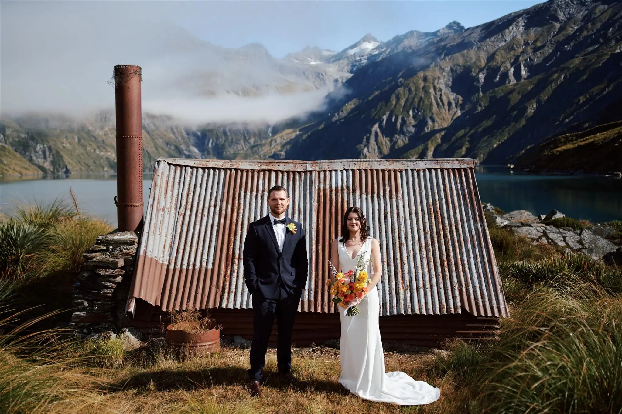 queenstown new zealand heli wedding elopement photographer videographer | A couple in formal attire stands in front of a rustic corrugated metal hut, with mountains and a lake in the background.