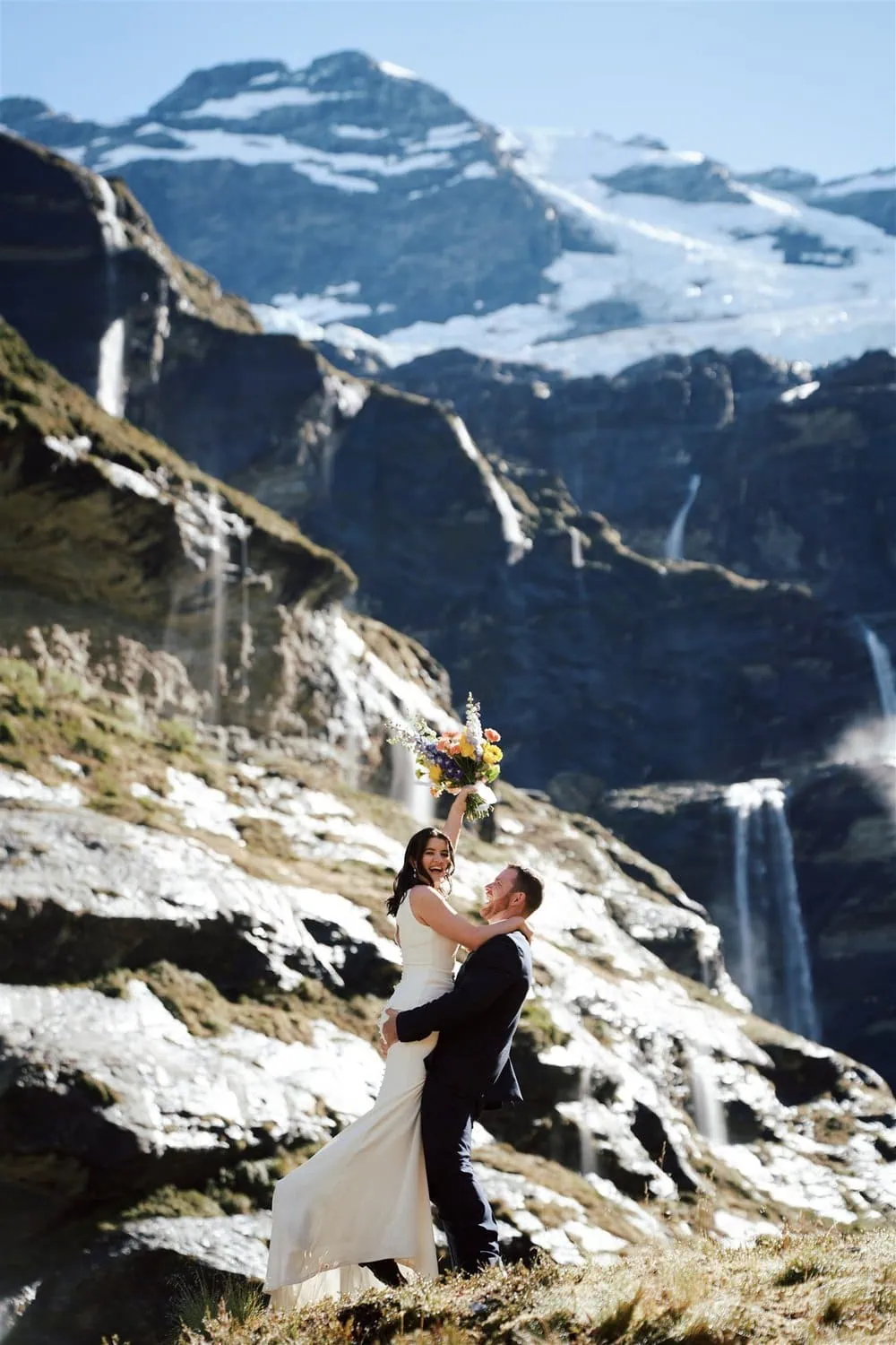 queenstown new zealand heli wedding elopement photographer videographer | A groom lifts a bride who holds a bouquet, set against a mountainous backdrop with waterfalls and a glacier.
