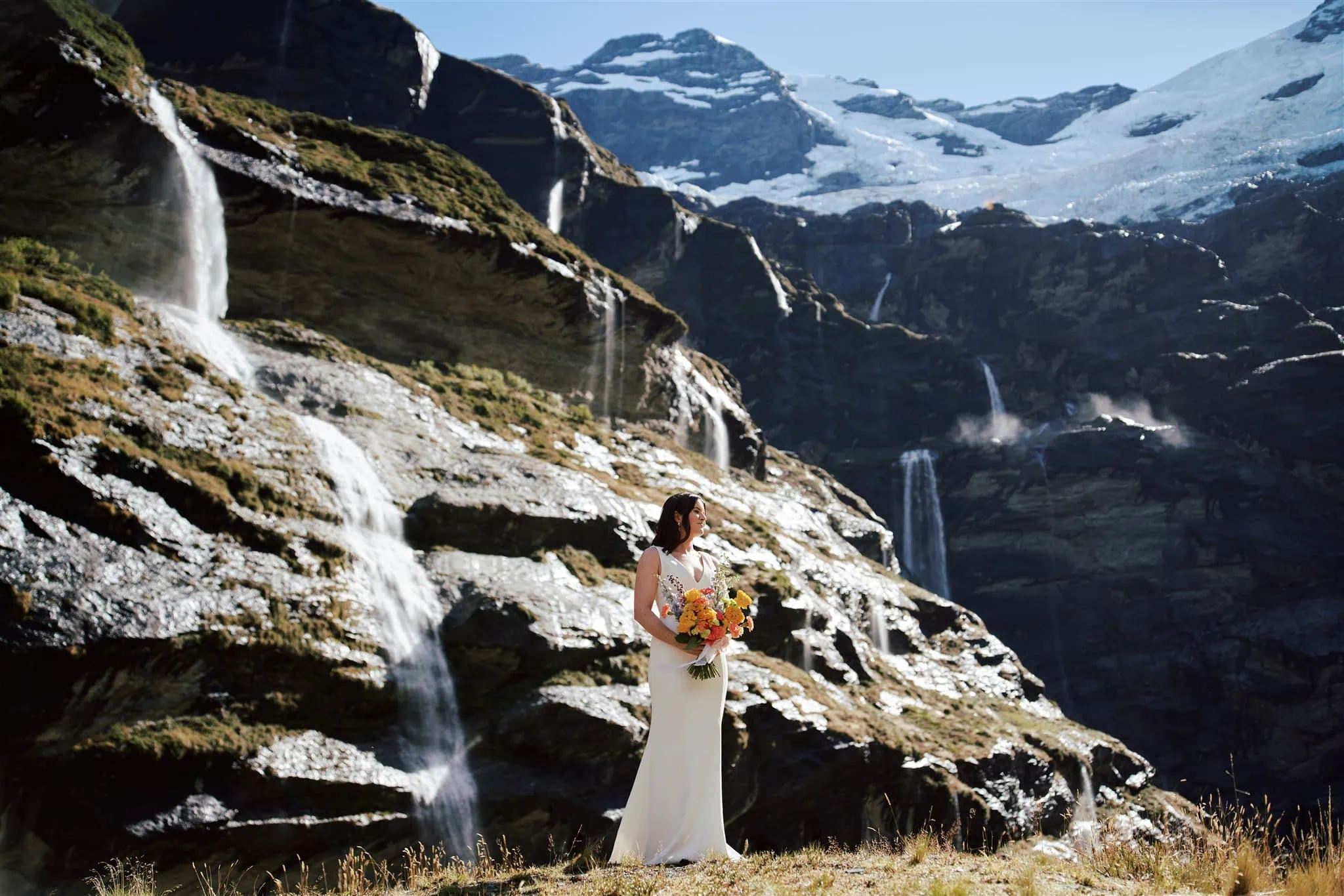 queenstown new zealand heli wedding elopement photographer videographer | A woman in a white dress holds a bouquet while standing in a mountainous landscape with multiple waterfalls.