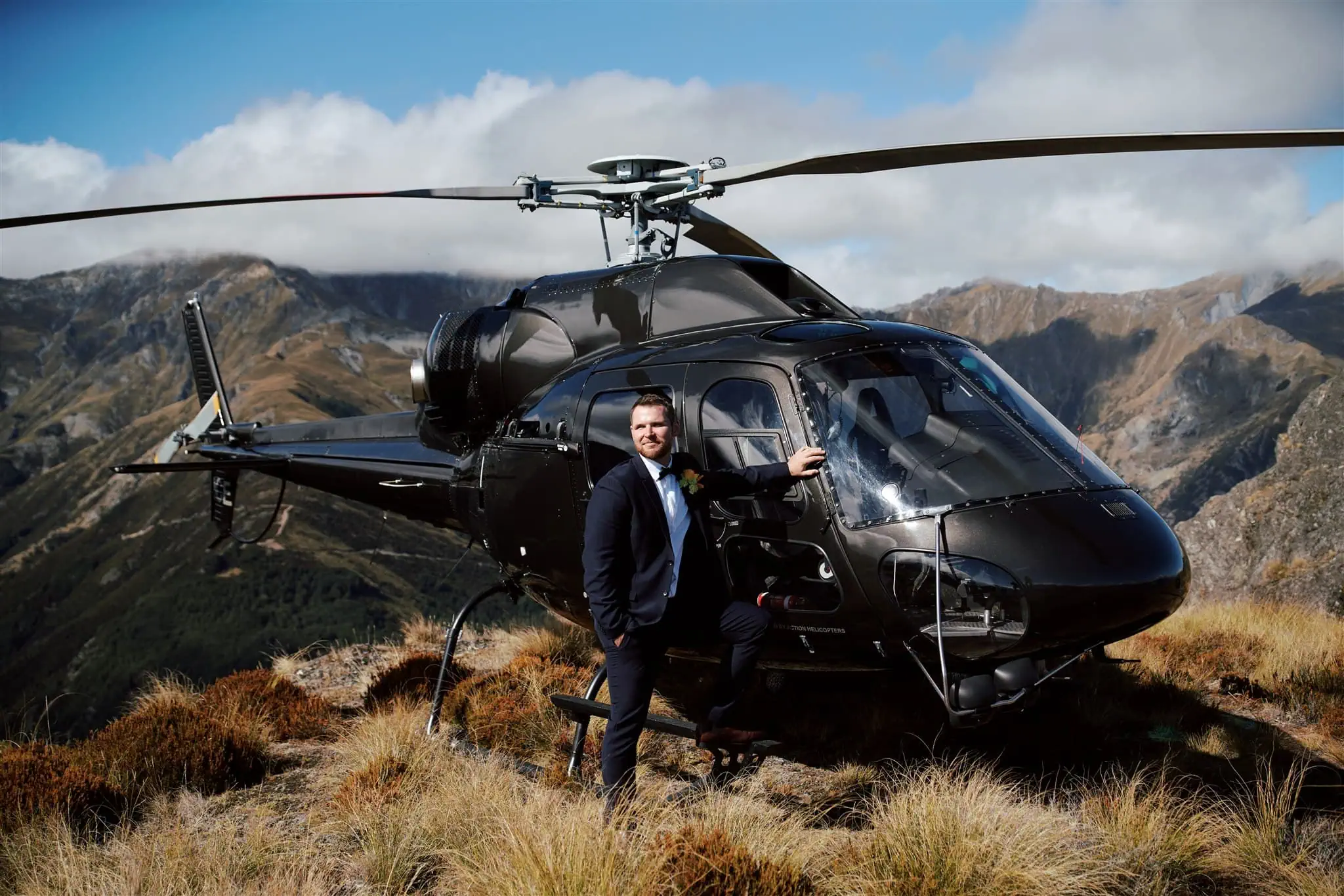 queenstown new zealand heli wedding elopement photographer videographer | A person in a suit stands next to a black helicopter on a grassy hilltop with mountains in the background.