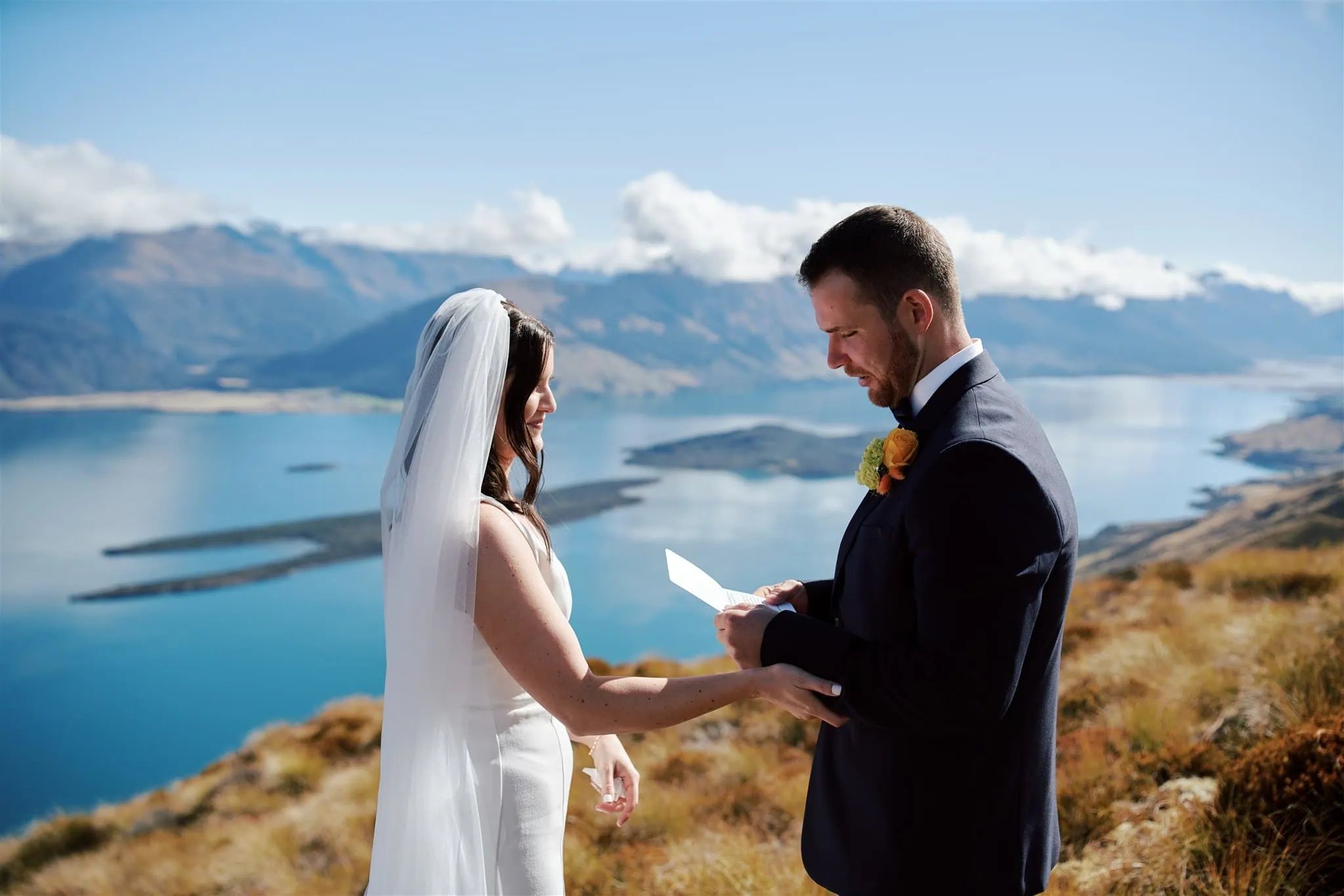 queenstown new zealand heli wedding elopement photographer videographer | A couple in wedding attire exchanges vows on a mountain overlooking a vast lake and mountain range under a clear blue sky.