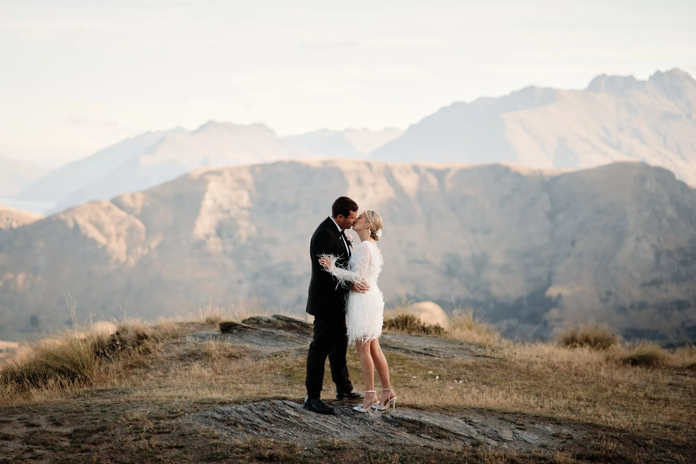 queenstown new zealand heli wedding elopement photographer videographer | A couple in formal attire embraces on a rocky hilltop with rugged mountains under a cloudy sky, savoring their Queenstown heli-wedding package's breathtaking moments.