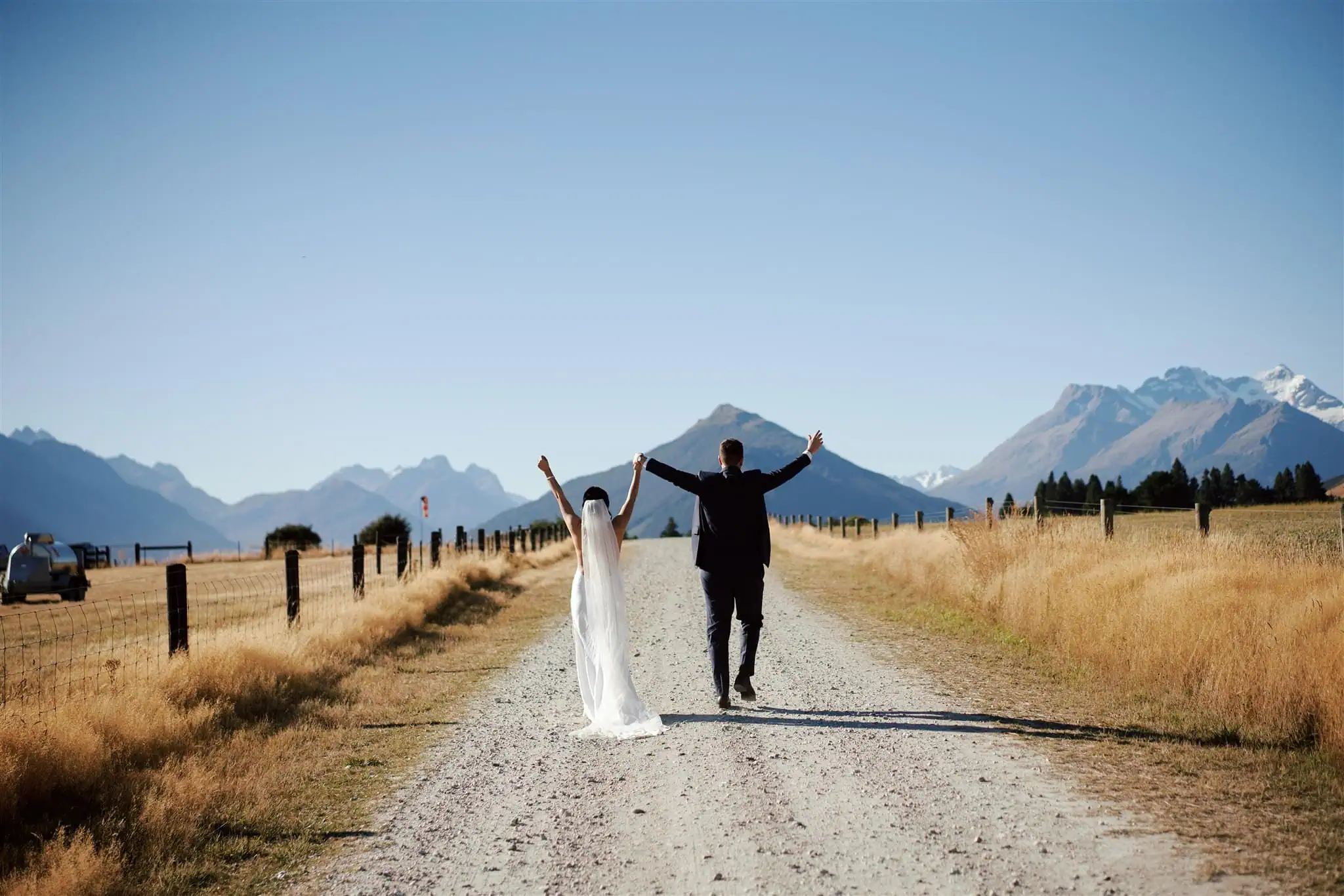 queenstown new zealand heli wedding elopement photographer videographer | A bride and groom walk away on a gravel road surrounded by fields and mountains, arms raised in celebration, as they savor their unforgettable Queenstown heli-wedding package.