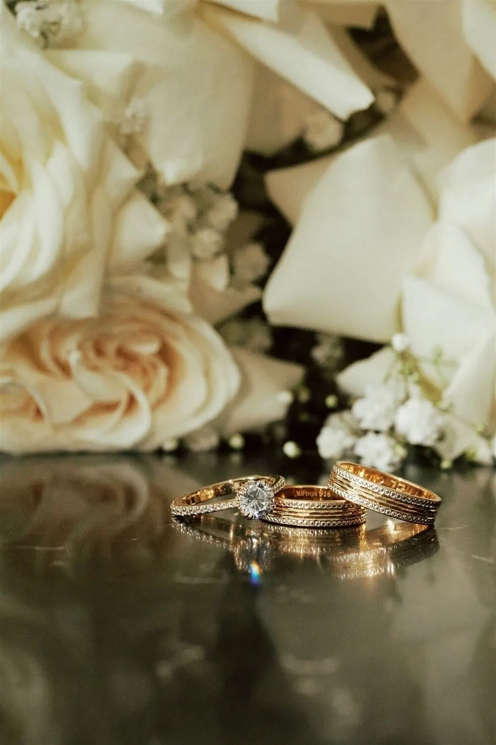 queenstown new zealand heli wedding elopement photographer videographer | Three gold rings, including a diamond engagement ring and two wedding bands, rest on a reflective surface at a Queenstown AirBnB, with white roses and baby's breath flowers in the background.