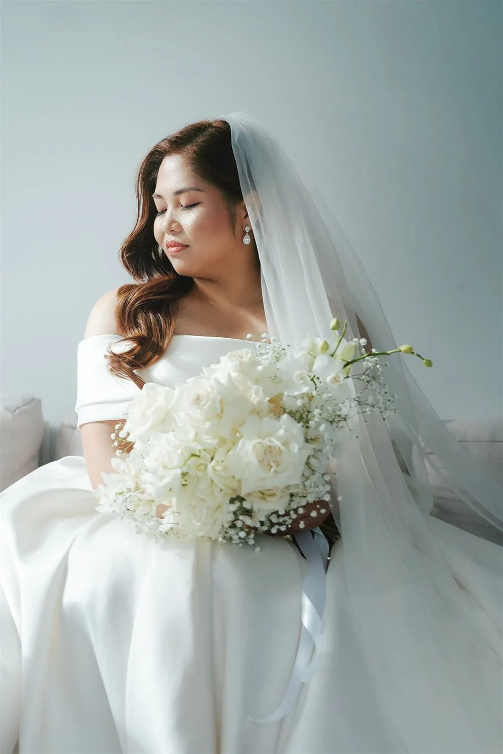 queenstown new zealand heli wedding elopement photographer videographer | A bride in a white dress and veil sits with her eyes closed, holding a bouquet of white roses and baby's breath against a light background at an elegant AirBnB near Jacks Point, Queenstown.