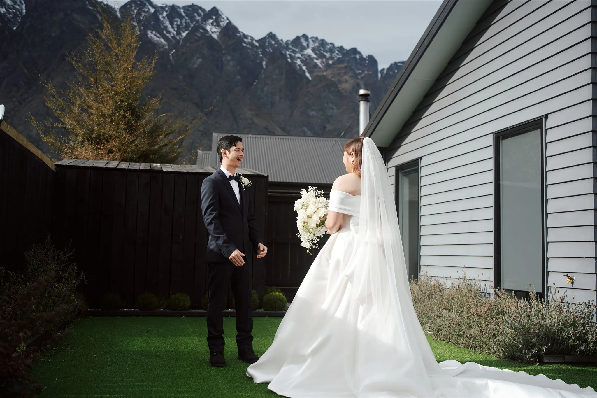 queenstown new zealand heli wedding elopement photographer videographer | A bride and groom stand facing each other outdoors at Jacks Point, Queenstown, with mountains in the background and a modern AirBnB building to the right. The bride holds a bouquet and wears a white dress and veil.