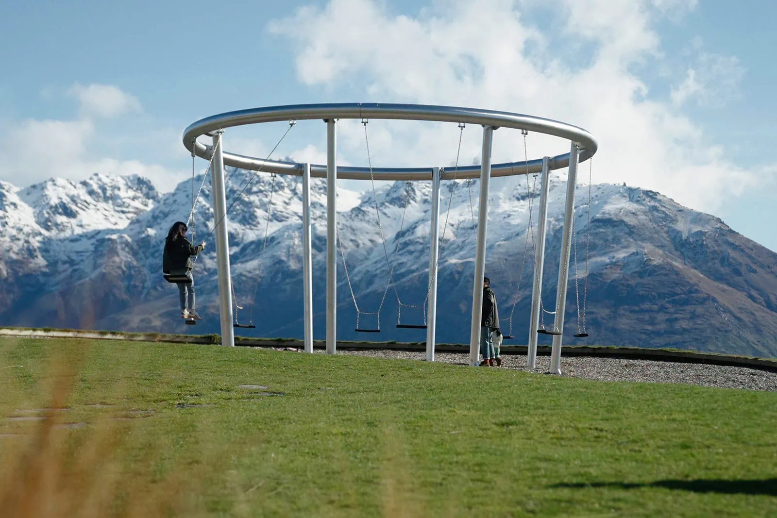 queenstown new zealand heli wedding elopement photographer videographer | A person sits on a swing attached to a circular metal frame, while another stands nearby at Jacks Point; snow-capped mountains rise in the background, promising an unforgettable Queenstown AirBnB short term accommodation experience.