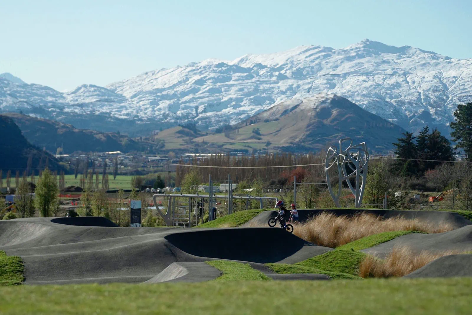 queenstown new zealand heli wedding elopement photographer videographer | A cyclist rides on a pump track in the foreground at Jacks Point, Queenstown, with grassy hills and snow-capped mountains in the background under a clear sky. Ideal for an adventurous AirBnB getaway.