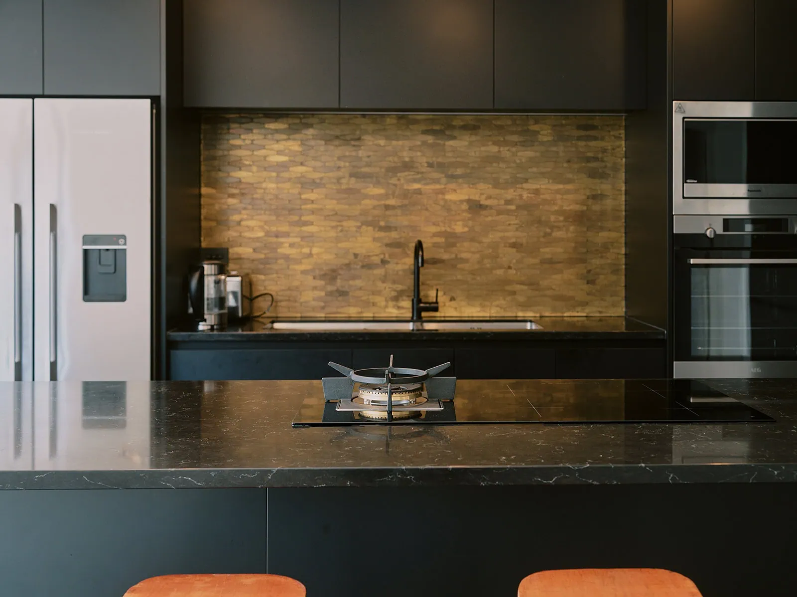 queenstown new zealand heli wedding elopement photographer videographer | Modern kitchen in Jacks Point with a black marble island, built-in gas burner, stainless steel refrigerator, oven, microwave, and a gold tile backsplash above the sink—perfect for your next Queenstown AirBnB stay.