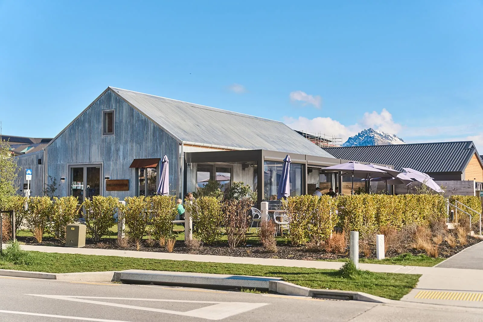 queenstown new zealand heli wedding elopement photographer videographer | A modern, metal-roofed café with outdoor seating and umbrellas is surrounded by shrubs on a sunny day beside a quiet street in Jacks Point, Queenstown.
