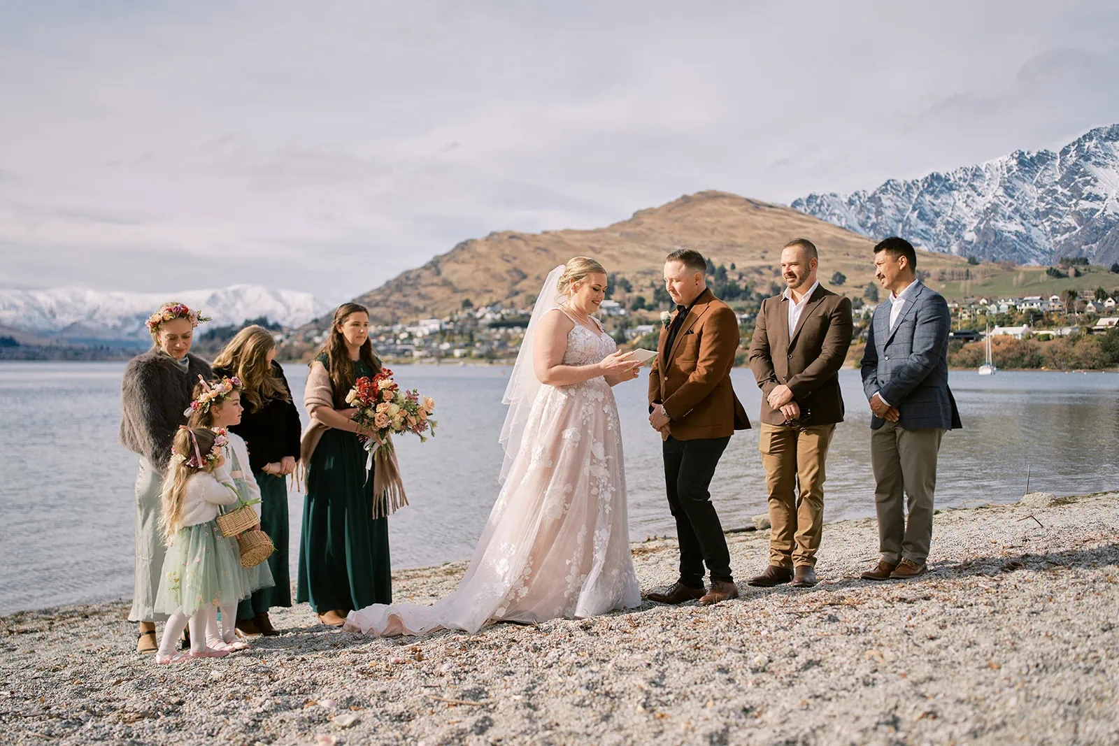 queenstown new zealand heli wedding elopement photographer videographer | A bride and groom stand facing each other on a lakeside beach at their Queenstown wedding, exchanging vows with their wedding party and flower girls, framed by majestic mountains and a charming town in the background.