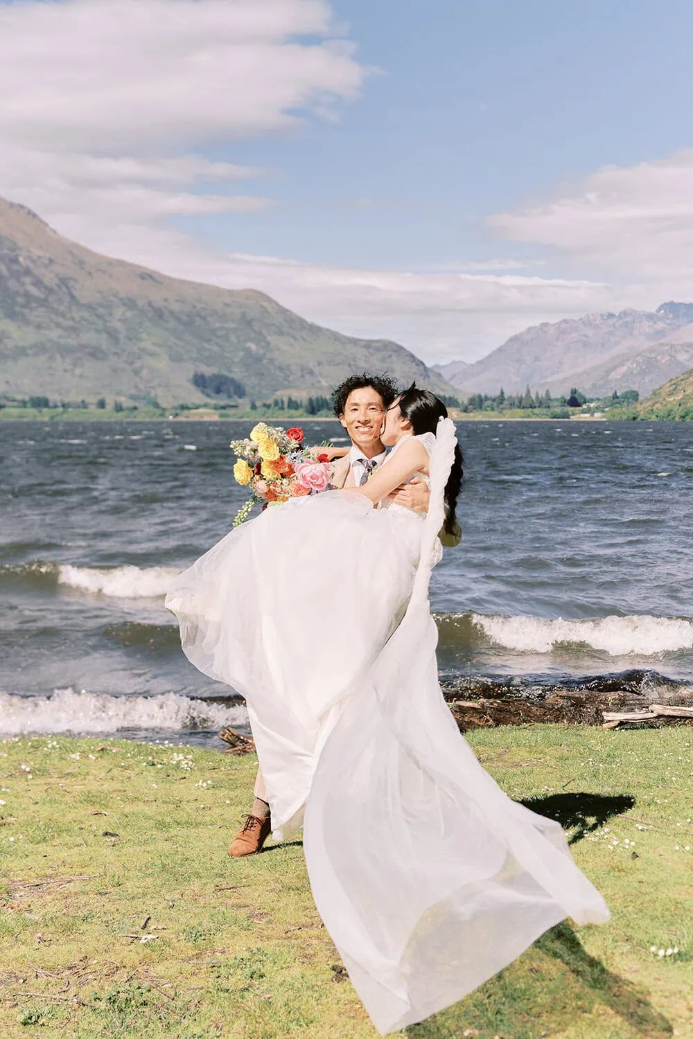 queenstown new zealand heli wedding elopement photographer videographer | A groom carries a bride in a white dress by the lakeside, mountains rising behind them under a partly cloudy sky—an unforgettable moment at Lake Hayes weddings.