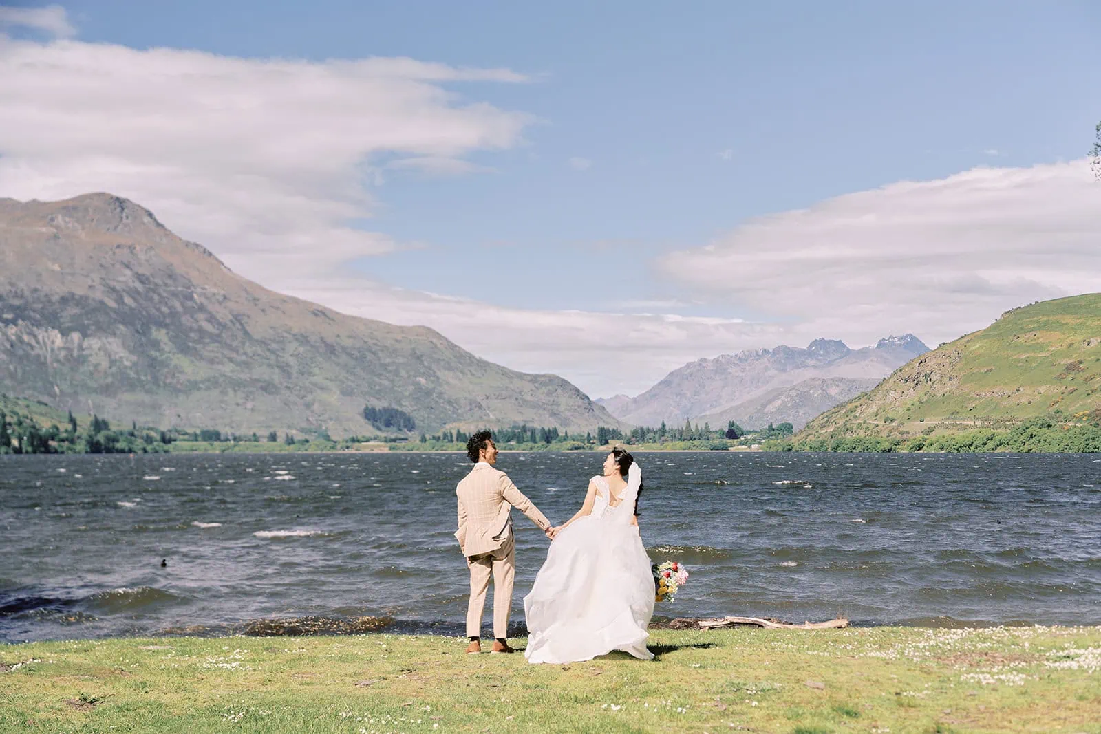 queenstown new zealand heli wedding elopement photographer videographer | A bride and groom stand hand-in-hand by the scenic Lake Hayes lakeshore, with mountains and blue sky in the background—an unforgettable setting for Queenstown weddings.