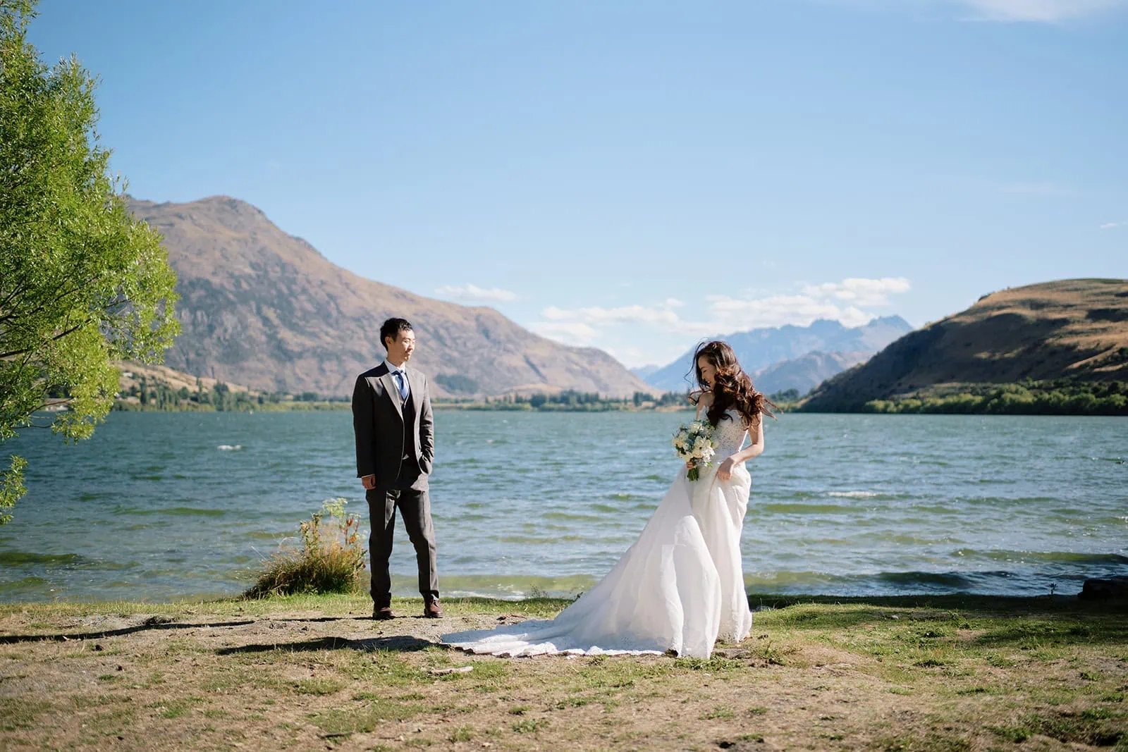 queenstown new zealand heli wedding elopement photographer videographer | A bride and groom stand by a lake with mountains in the background on a sunny day; perfect for Lake Hayes weddings, the bride holds a bouquet and looks down at her dress while the groom faces her.