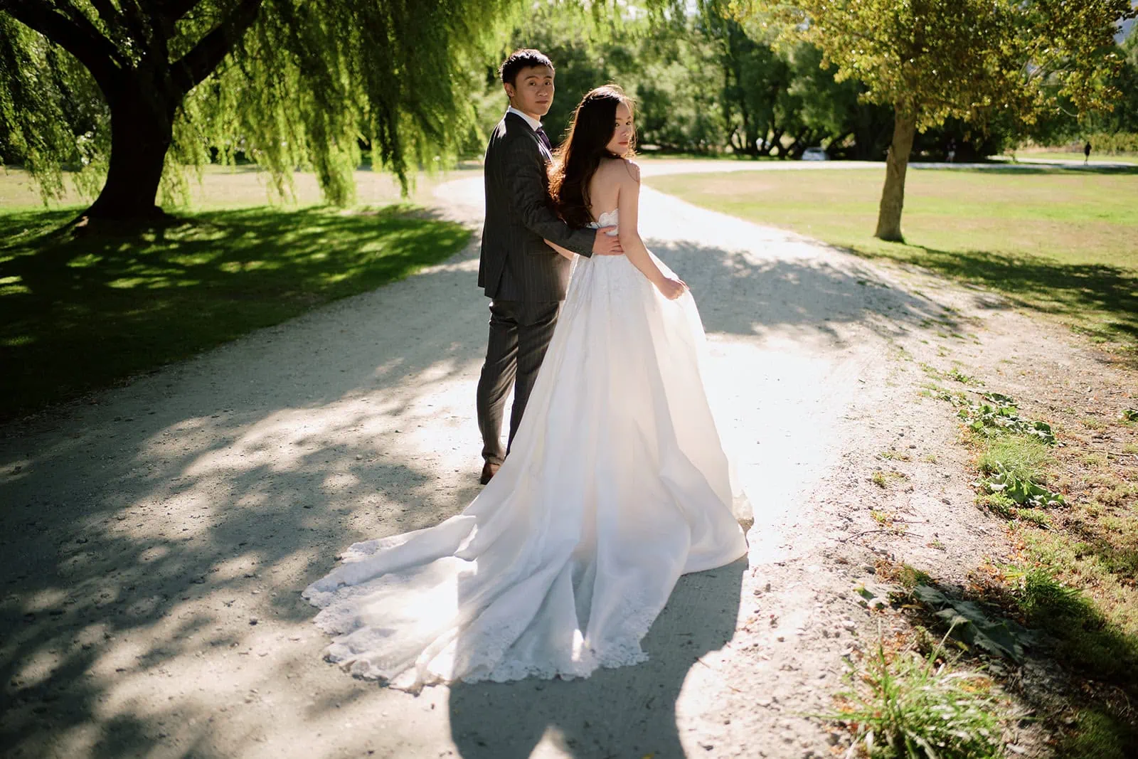 queenstown new zealand heli wedding elopement photographer videographer | A bride and groom stand on a sunlit path in a park, surrounded by trees and grass. Perfect for Queenstown weddings, the bride’s long white gown and the groom’s suit capture the elegance of this unforgettable day.