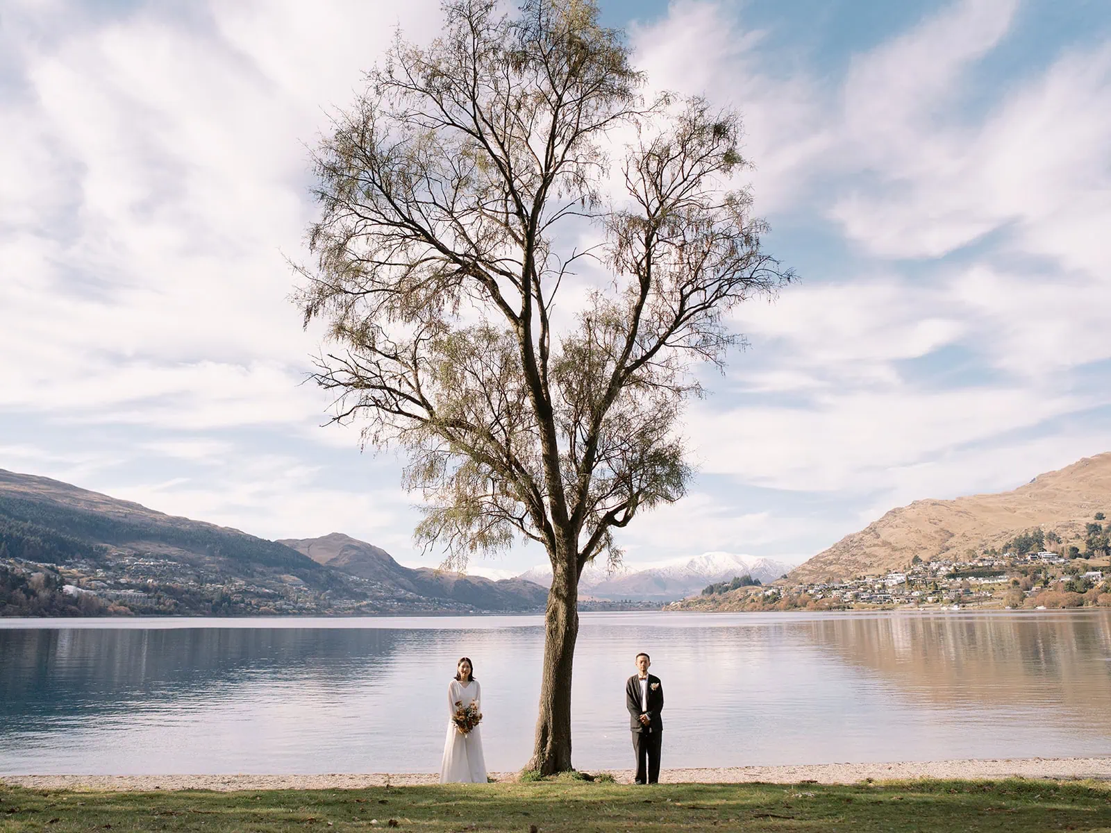 queenstown new zealand heli wedding elopement photographer videographer | A bride and groom stand on opposite sides of a tree by a lake, with mountains and a partly cloudy sky in the background—an idyllic setting for Queenstown weddings.