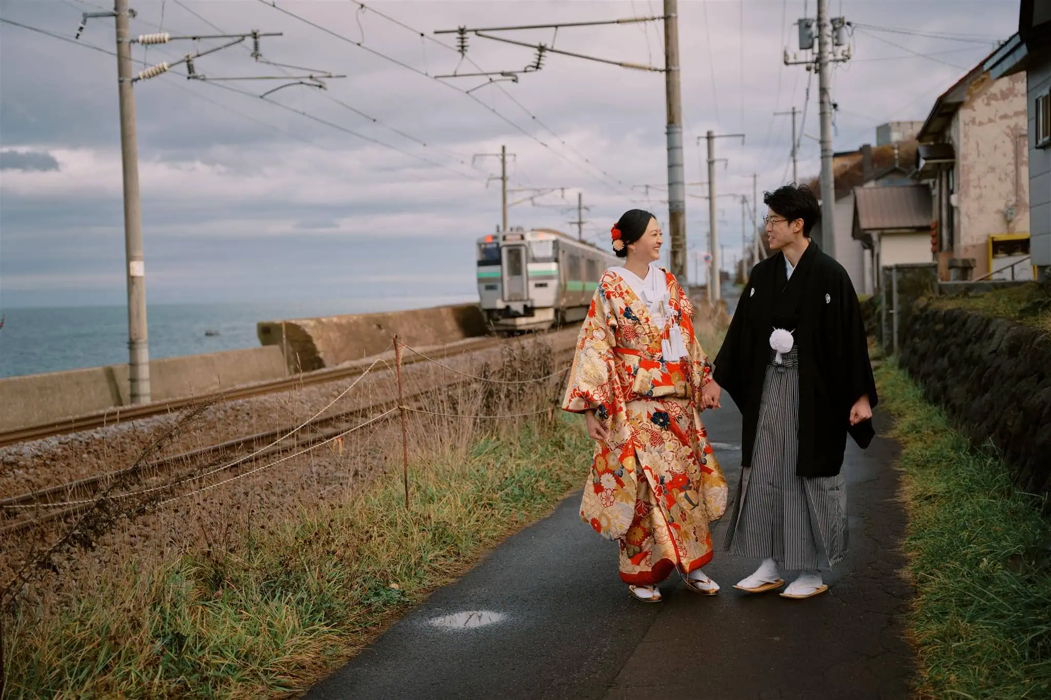 queenstown new zealand heli wajapan elopement packages nomad weddings kyoto tokyo nisekoedding elopement photographer videographer | A couple wearing traditional Japanese clothing walks on a seaside path near railway tracks as a train passes by—capturing the romantic spirit of eloping in Japan, far from the bustle of Kyoto’s popular wedding packages.